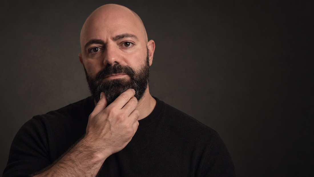North Common founder with a full dark beard, wearing a black shirt and gently touching his beard against a dark charcoal studio background.
